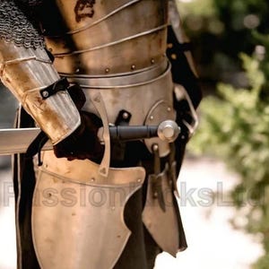 Puede incluir: Primer plano de una armadura medieval, que incluye una coraza de metal, protectores de brazos y cota de malla. Una espada con una hoja plateada y un mango negro se sostiene en una mano enguantada. La armadura tiene una apariencia desgastada.