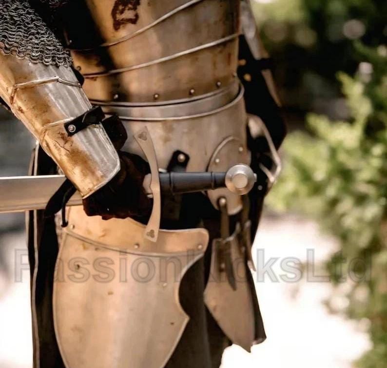 Puede incluir: Primer plano de una armadura medieval, que incluye una coraza de metal, protectores de brazos y cota de malla. Una espada con una hoja plateada y un mango negro se sostiene en una mano enguantada. La armadura tiene una apariencia desgastada.