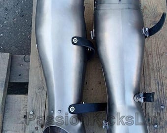 May include: A pair of polished metal greaves, possibly for armour, with curved forms and black leather straps. The greaves are displayed on a wooden surface. The metal has a reflective quality, suggesting a smooth finish.