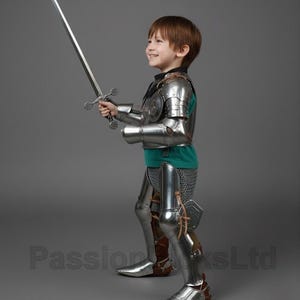 May include: A child in a full suit of shiny, silver-colored knight's armor, including a breastplate, arm and leg guards, and boots. The child is holding a long, silver sword with a detailed hilt. The child is wearing a green shirt under the armor.