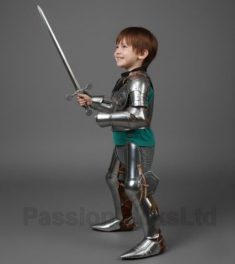 May include: A child in a full suit of shiny, silver-colored knight's armor, including a breastplate, arm and leg guards, and boots. The child is holding a long, silver sword with a detailed hilt. The child is wearing a green shirt under the armor.