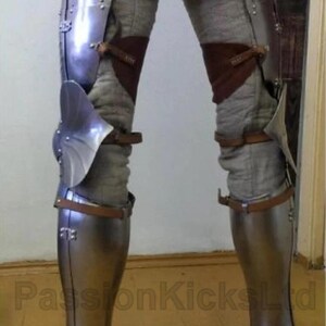 May include: Medieval leg armor made of polished steel, featuring articulated plates and leather straps. The armor covers the thighs, knees, and shins, with a foot section. The legs are covered in a gray fabric. The armor is designed for protection.