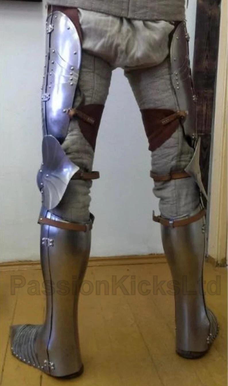 May include: Medieval leg armor made of polished steel, featuring articulated plates and leather straps. The armor covers the thighs, knees, and shins, with a foot section. The legs are covered in a gray fabric. The armor is designed for protection.