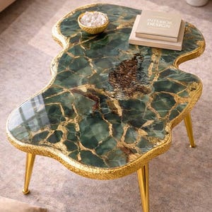 Könnte beinhalten: Einzigartiger Couchtisch mit grün-goldener Musterplatte. Der Tisch hat eine freie Form, goldene Verzierungen und goldene Beine. Eine kleine goldene Schale und Bücher befinden sich auf dem Tisch. Der Tisch ist ca. 76 cm lang.