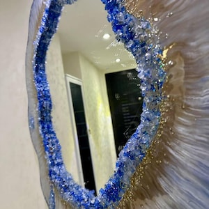 Puede incluir: Espejo de pared ovalado con un diseño único. El marco del espejo presenta un borde azul y transparente similar a un cristal, con un borde exterior blanco y dorado similar a la resina. El espejo refleja una habitación con paredes blancas y una puerta negra.