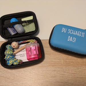 Könnte beinhalten: Zwei kleine schwarze Reißverschlussetuis, eines offen und eines geschlossen, auf einer Holzoberfläche. Das offene Etui enthält eine Vielzahl kleiner Gegenstände, darunter ein Spitzer, ein Holzspielzeug und ein kleiner Notizblock. Das geschlossene Etui ist blau mit dem Text "DU SCHAFFST DAS!"