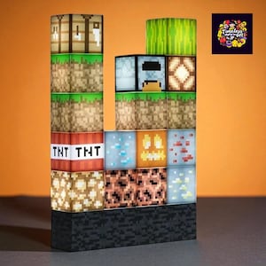 May include: A Minecraft-themed lamp composed of various illuminated blocks, including TNT, dirt, and a watermelon block. The lamp is stacked in a tower formation, with a black base and an orange background. The lamp has a logo in the upper right corner.