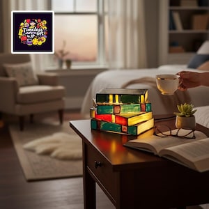 May include: A stack of colourful, illuminated book-shaped lamps on a dark wooden bedside table. The lamps are green, red, and yellow. A cup of tea, a succulent, and an open book with glasses are also on the table. The image includes the text "Timeless Gift by JP".