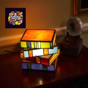 May include: A stack of stained glass book-shaped lamps in shades of orange, yellow, blue, and red. The lamps are illuminated, casting a warm glow. A vintage-style record player sits nearby. The image includes a logo that says "Timeless Gift by JP."