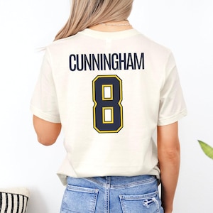 May include: A cream-colored short-sleeve t-shirt featuring the name CUNNINGHAM printed in dark blue capital letters above a large dark blue number 8 with a yellow outline on the back of the garment, paired with blue denim jeans.