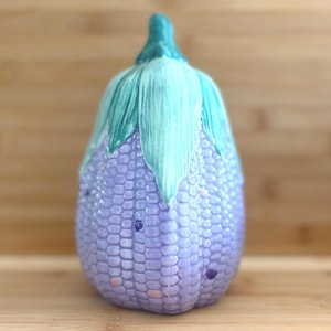 May include: A ceramic corn-shaped ornament. The body is a light purple with a textured pattern, and the top features green and blue-green leaves. It is set against a wooden background.