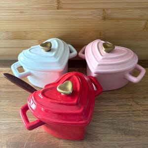 May include: Three heart-shaped ceramic ramekins in white, pink, and red, each with a gold-colored heart-shaped knob on the lid and small handles. A wooden spoon rests in the red ramekin. The ramekins are on a wooden surface.