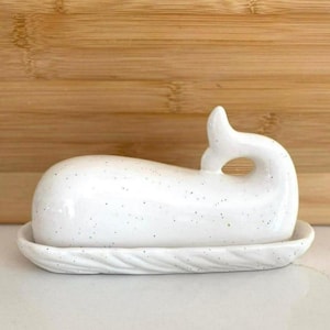 May include: A white ceramic butter dish in the shape of a whale. The dish has a speckled finish and sits on a matching base with a textured, wave-like design. The whale's tail forms a handle.