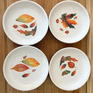 May include: Four white ceramic plates with speckled patterns and colorful autumn leaf designs. Each plate features a unique arrangement of red, orange, and green leaves, perfect for serving appetizers or desserts.