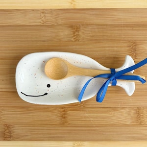 May include: A white ceramic whale-shaped spoon rest with a small wooden spoon. The spoon rest has a speckled design and a blue ribbon tied around the spoon and the whale's tail. The whale has a smiling face.