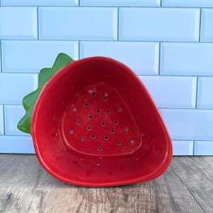 May include: A ceramic strawberry-shaped colander. The colander is bright red with small drainage holes and a green leafy top. It sits on a wooden surface against a light blue tiled background. This kitchenware item is designed for washing and draining fruits.