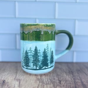 May include: A ceramic mug with a forest scene design. The mug features a green handle and a two-tone design with a green and white color scheme. The bottom half of the mug has a forest silhouette design.