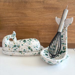 May include: A ceramic butter dish and whisk holder, both whale-shaped, with a white base and green and gold speckled pattern. A stainless steel whisk rests in the holder. The butter dish has a lid and sits on a matching plate.