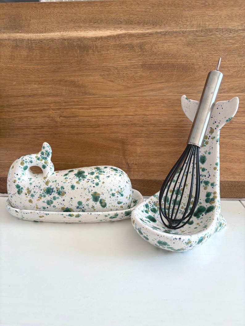 May include: A ceramic butter dish and whisk holder, both whale-shaped, with a white base and green and gold speckled pattern. A stainless steel whisk rests in the holder. The butter dish has a lid and sits on a matching plate.
