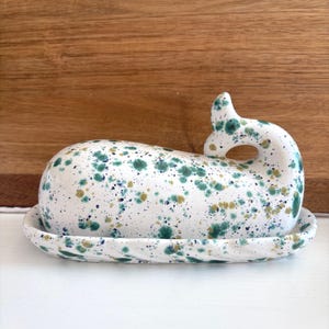May include: A ceramic butter dish in the shape of a whale, with a white base and lid. The dish is speckled with green, blue, and gold paint. The whale's tail forms a handle. The dish sits on a white surface.