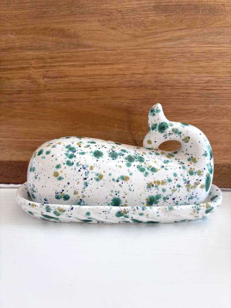 May include: A ceramic butter dish in the shape of a whale, with a white base and lid. The dish is speckled with green, blue, and gold paint. The whale's tail forms a handle. The dish sits on a white surface.
