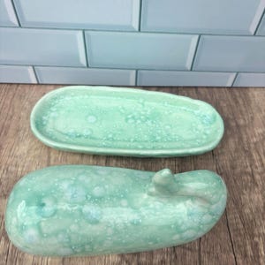 May include: Two ceramic pieces, a long oval dish and a rounded, oblong object, both in a speckled, seafoam green glaze. The dish is shallow, while the oblong piece has a small, raised detail on one end. The background is a light blue tile.