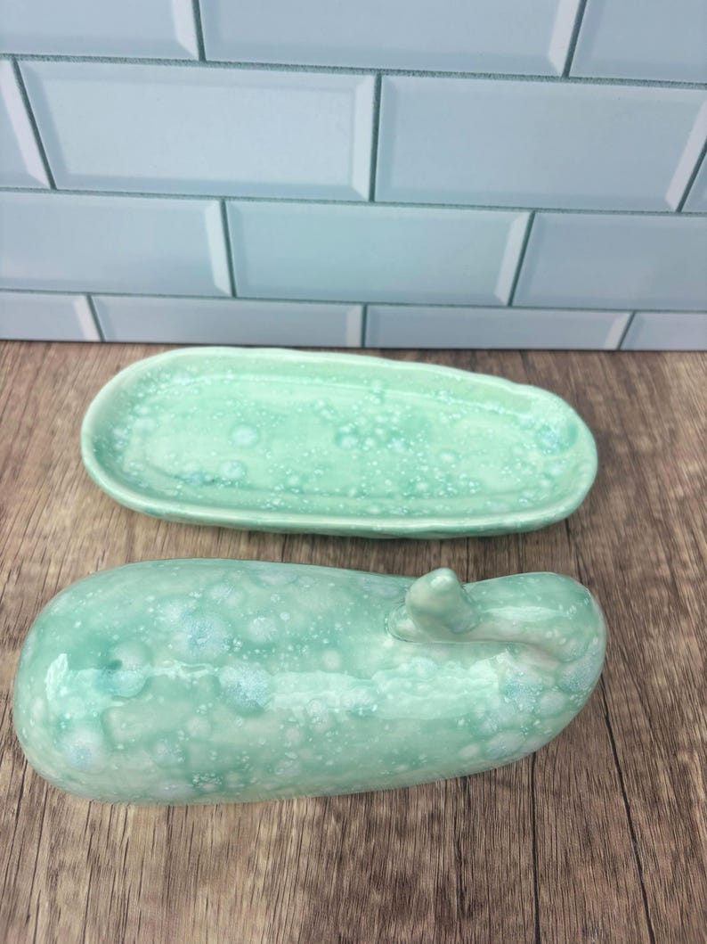 May include: Two ceramic pieces, a long oval dish and a rounded, oblong object, both in a speckled, seafoam green glaze. The dish is shallow, while the oblong piece has a small, raised detail on one end. The background is a light blue tile.