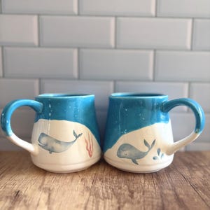 May include: Two ceramic mugs with a blue and white design. Each mug features a blue whale illustration and a blue handle. The mugs have a glossy finish and are set against a light blue tile background.