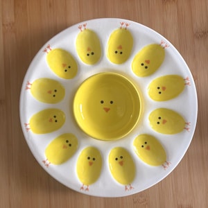 May include: A white ceramic serving platter with a yellow center bowl and egg-shaped compartments. Each compartment is decorated with a yellow chick design, featuring black eyes and orange feet. The platter is ideal for serving snacks or appetizers.