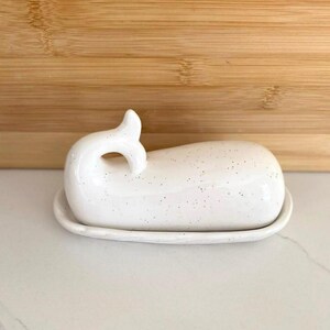 May include: A white ceramic butter dish in the shape of a whale. The dish has a speckled pattern and a curved tail handle. The base is oval-shaped, and the dish is sitting on a white surface.