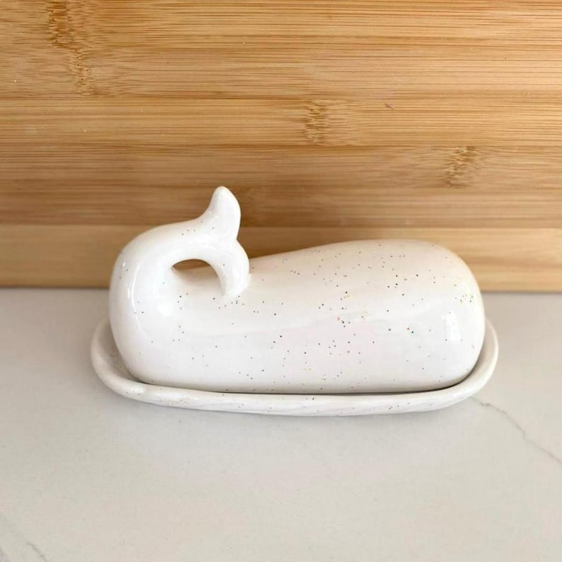 May include: A white ceramic butter dish in the shape of a whale. The dish has a speckled pattern and a curved tail handle. The base is oval-shaped, and the dish is sitting on a white surface.