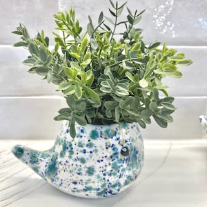 May include: A white ceramic planter shaped like a whale with blue and green splatter paint. The planter is filled with a green leafy plant.