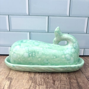 May include: A seafoam green ceramic butter dish in the shape of a whale. The dish has a textured surface with white speckles and sits on a matching oval base. The whale's tail forms a handle.