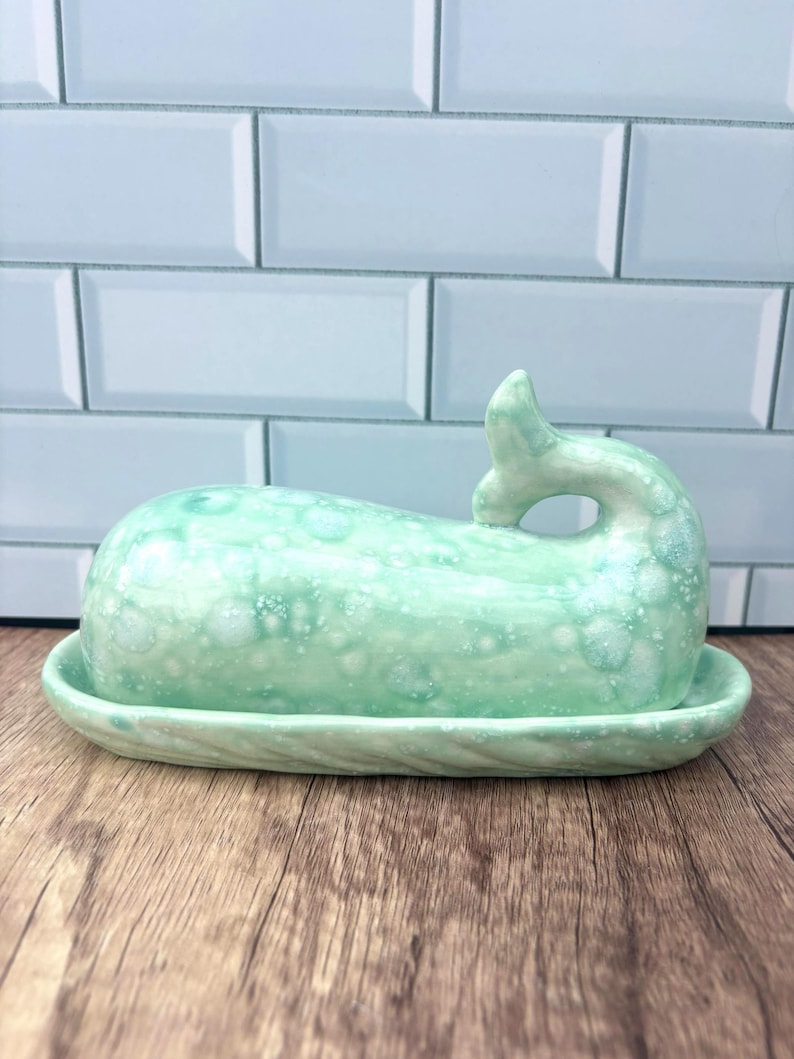 May include: A seafoam green ceramic butter dish in the shape of a whale. The dish has a textured surface with white speckles and sits on a matching oval base. The whale's tail forms a handle.