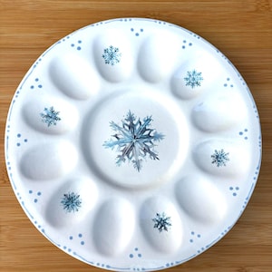 May include: White ceramic serving plate with a snowflake design. The plate features a central snowflake and smaller snowflakes around the edges. The rim has a dotted blue border. The plate has 12 oval-shaped compartments.