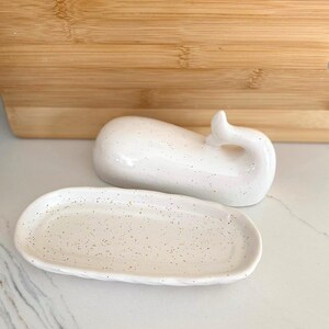 May include: A white ceramic whale-shaped trinket dish with a matching oval tray. Both pieces have a speckled design. The whale has a curved tail. The tray is oval-shaped with a slightly raised edge. The background includes a wooden cutting board.
