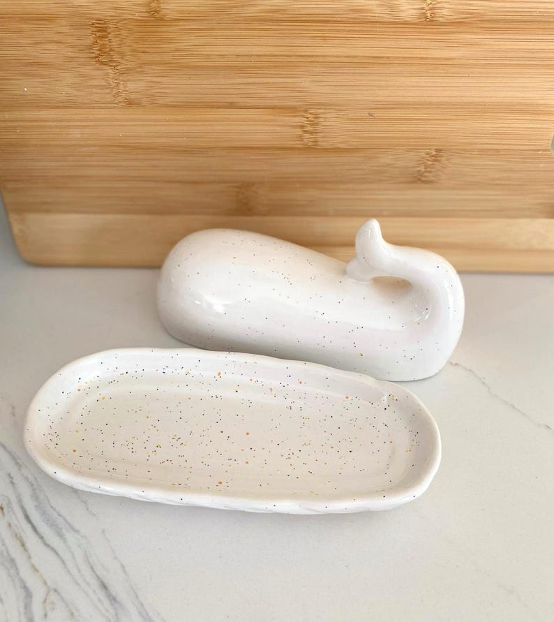 May include: A white ceramic whale-shaped trinket dish with a matching oval tray. Both pieces have a speckled design. The whale has a curved tail. The tray is oval-shaped with a slightly raised edge. The background includes a wooden cutting board.