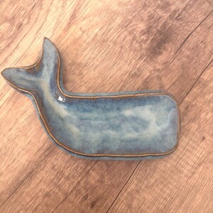 May include: A ceramic whale-shaped dish featuring a glossy blue glaze with mottled light and dark tones. The edges of the whale are defined by a distinct, thin brown outline that follows the contour of the tail and the elongated, rectangular body.