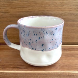 May include: A handmade ceramic mug with a white base and a lavender top speckled with dark blue dots. The mug has a curved handle and is set against a wooden surface. This unique mug is perfect for coffee, tea, or any beverage.