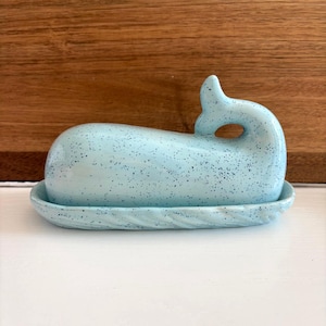 May include: A light blue ceramic whale-shaped butter dish with a speckled pattern. The dish includes a base with a textured rim and a whale-shaped lid with a curved tail and a blowhole. The dish is designed to hold butter.
