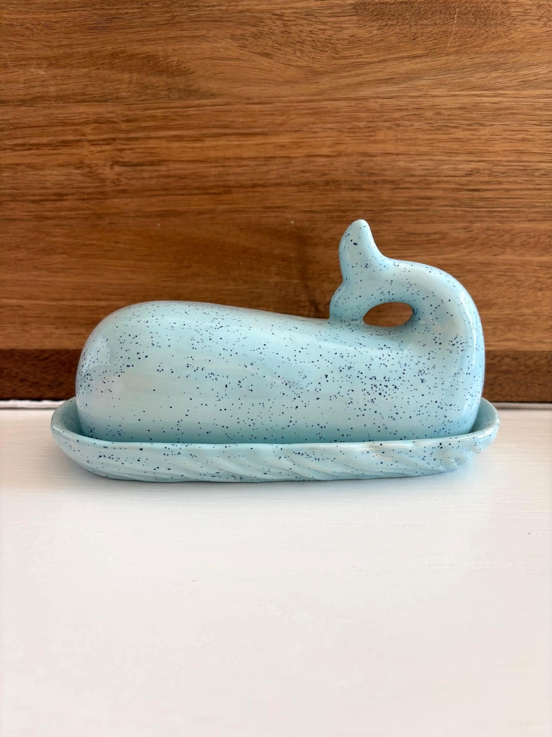 May include: A light blue ceramic whale-shaped butter dish with a speckled pattern. The dish includes a base with a textured rim and a whale-shaped lid with a curved tail and a blowhole. The dish is designed to hold butter.
