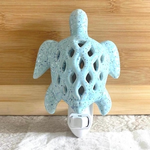 May include: A light blue ceramic night light shaped like a sea turtle. The turtle has a speckled pattern and diamond-shaped cutouts on its shell. The night light plugs directly into a wall outlet.