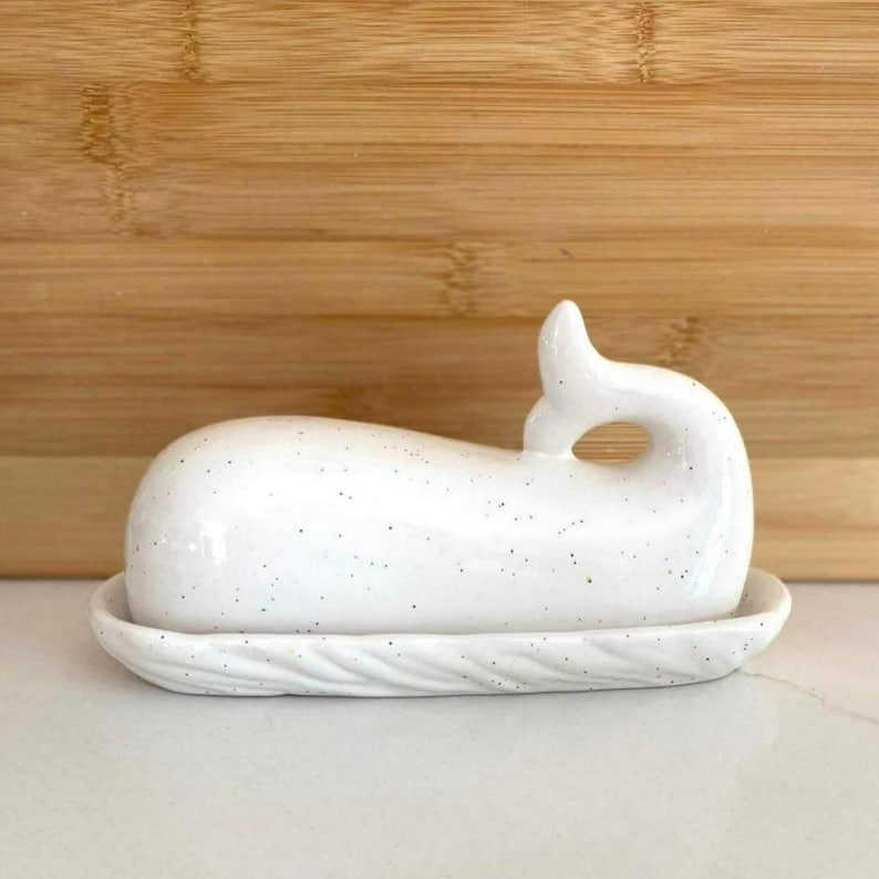 May include: A white ceramic butter dish in the shape of a whale, with a speckled finish. The dish has a curved body and a tail fin handle. The base is oval with a textured, rope-like border. The background is a wooden surface.