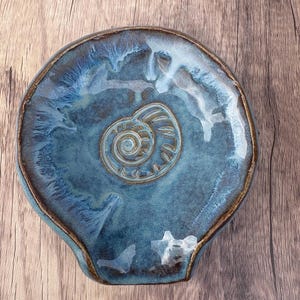 May include: A ceramic spoon rest with a glossy blue and white glaze featuring a raised spiral shell motif in the center. The dish has an irregular, rounded shape with a dark brown rim and a smooth, reflective surface finish throughout the piece.