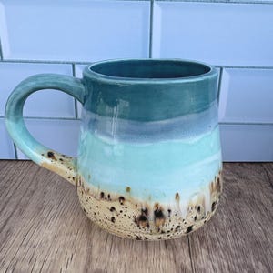 May include: A handmade ceramic mug with a unique glaze. The mug features a gradient of colors, including teal, light blue, and beige with brown speckles. The handle is teal. The mug is on a wooden surface.