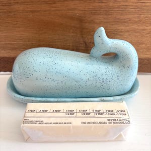 May include: A light blue ceramic whale-shaped butter dish with a matching base. The dish has a speckled design and a curved tail. A stick of butter is in front of the dish. The butter is 4 oz (113 g).