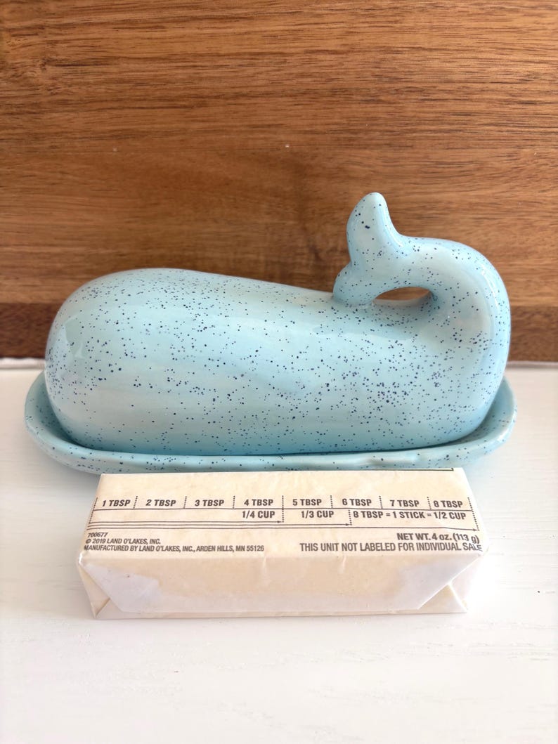 May include: A light blue ceramic whale-shaped butter dish with a matching base. The dish has a speckled design and a curved tail. A stick of butter is in front of the dish. The butter is 4 oz (113 g).