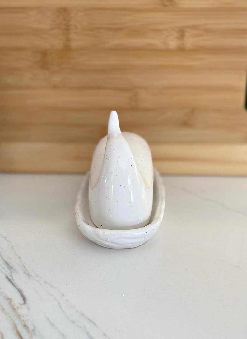 May include: A white ceramic garlic keeper with a speckled finish, resting on a matching oval dish. The garlic keeper has a rounded body and a pointed top. The set is designed for kitchen use.