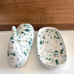 May include: Two ceramic pieces with a white base and a speckled design in shades of green and gold. One piece is a whale-shaped soap dish, and the other is an oval-shaped dish.