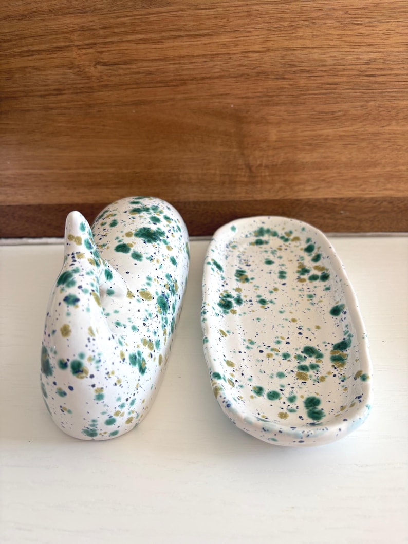 May include: Two ceramic pieces with a white base and a speckled design in shades of green and gold. One piece is a whale-shaped soap dish, and the other is an oval-shaped dish.